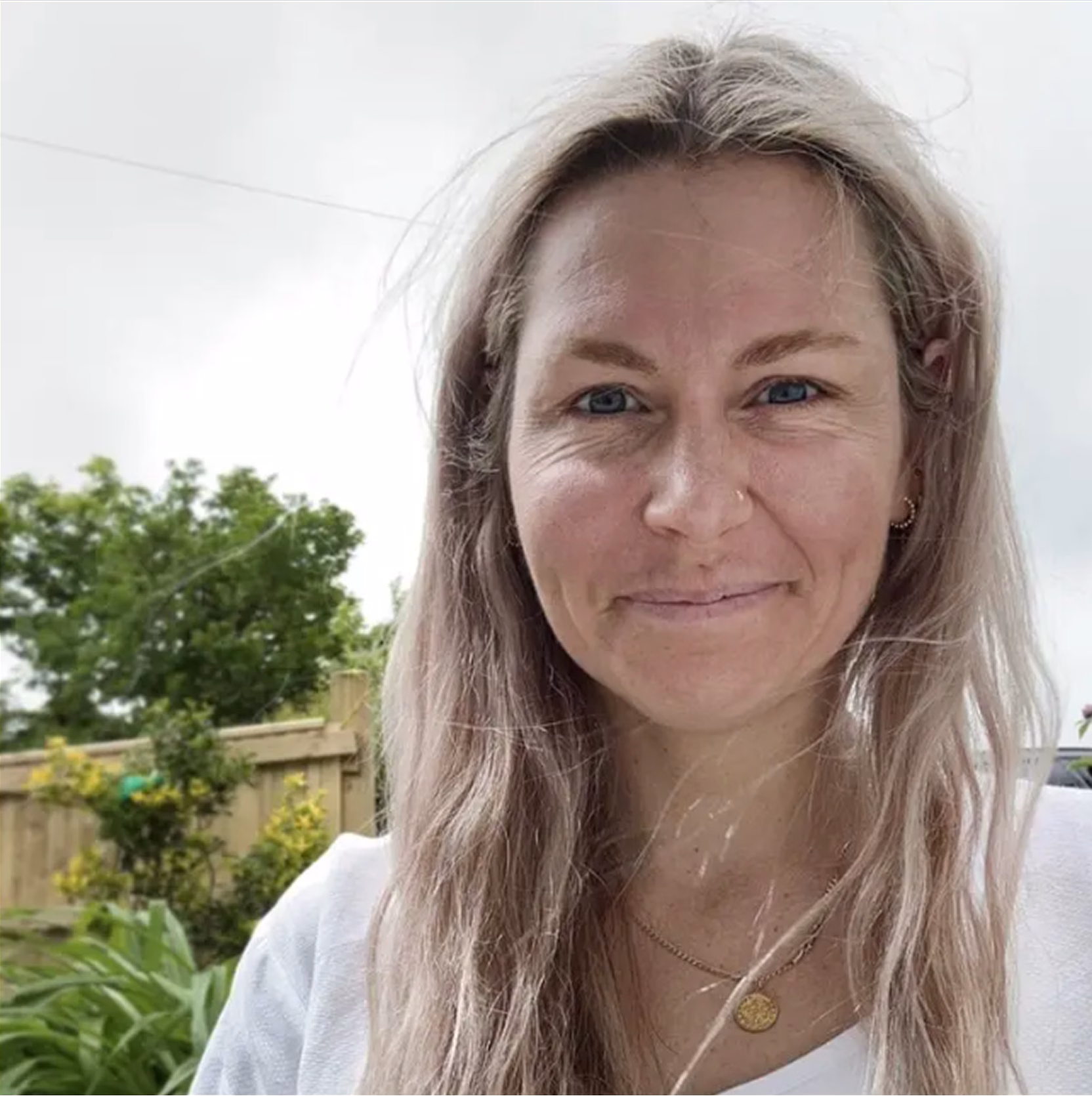 A smiling person with long, light-coloured hair wears a white top and a gold necklace with a circular pendant. They stand outdoors in front of a wooden fence, surrounded by green plants and trees under a cloudy sky. The natural setting and relaxed expression evoke a warm, approachable mood.
