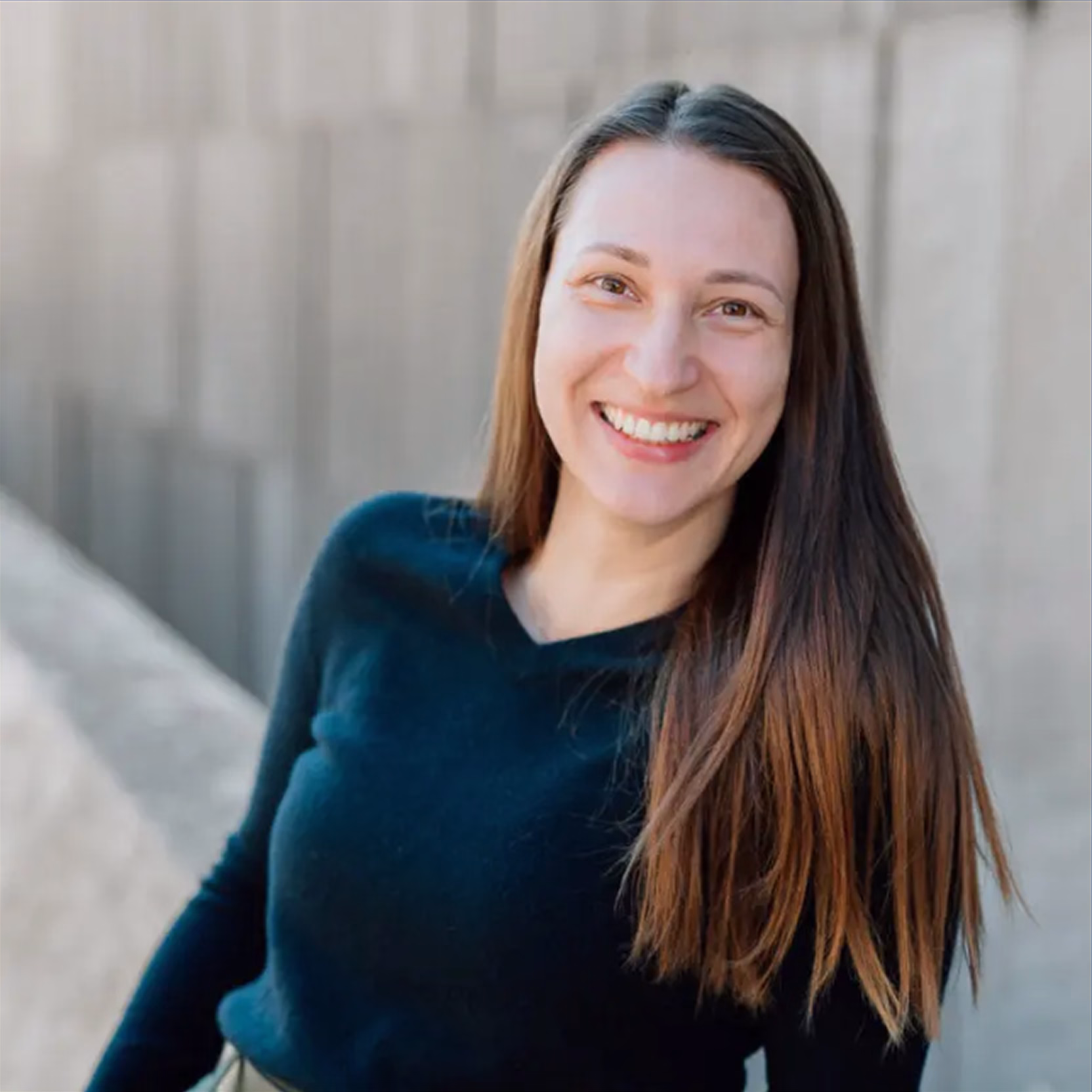 A smiling person with long straight brown hair stands outdoors against a textured concrete wall, wearing a dark long-sleeve top. Bright natural light highlights their relaxed expression, creating a calm and approachable mood.