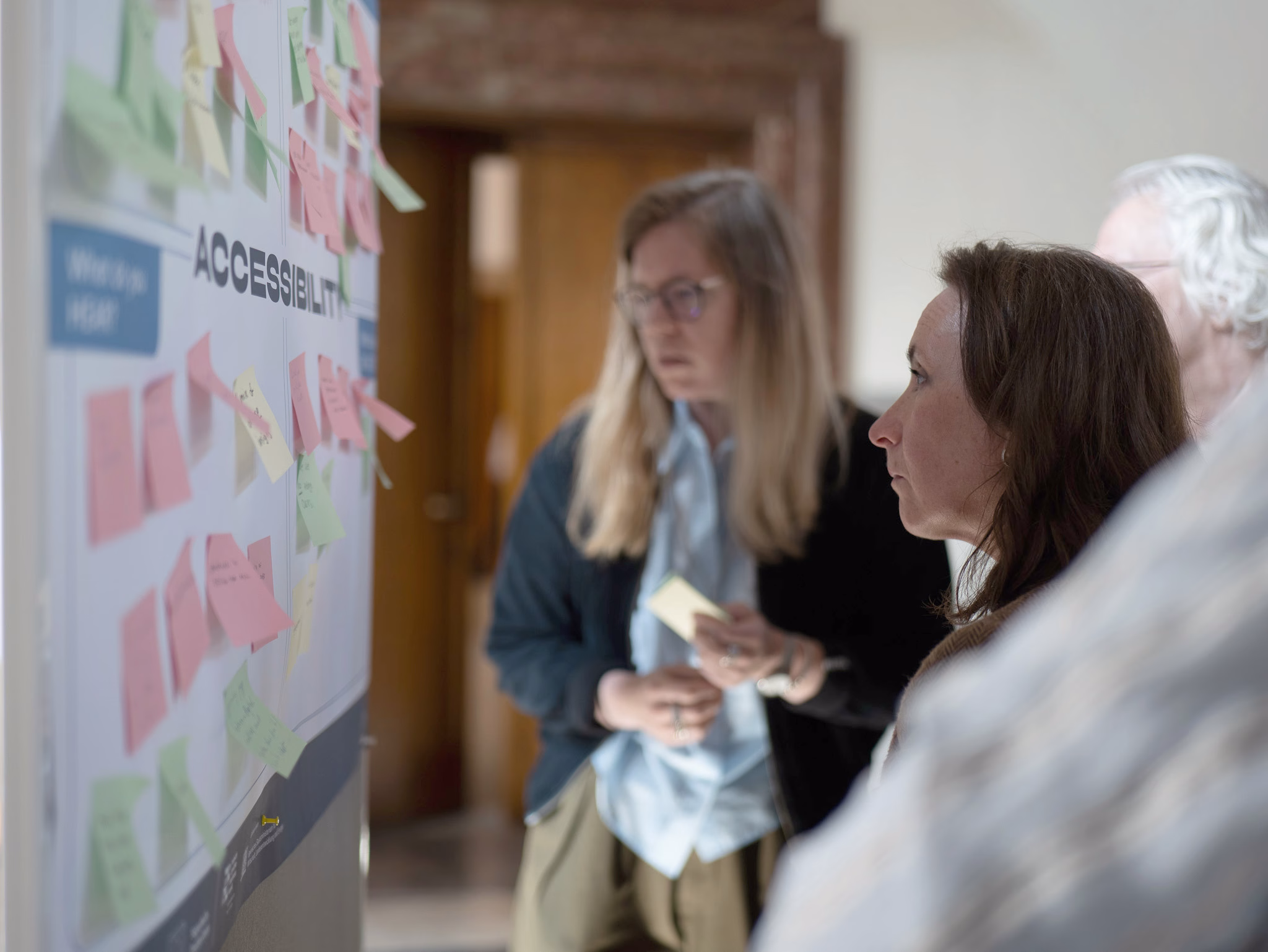 Participants review and discuss ideas on an accessibility workshop board covered with colourful sticky notes. Several people stand in front of the board, reading and contributing feedback, while the heading “ACCESSIBILITY” is visible among the posted notes. The image captures a collaborative co-creation session focused on sharing insights and developing ideas.
