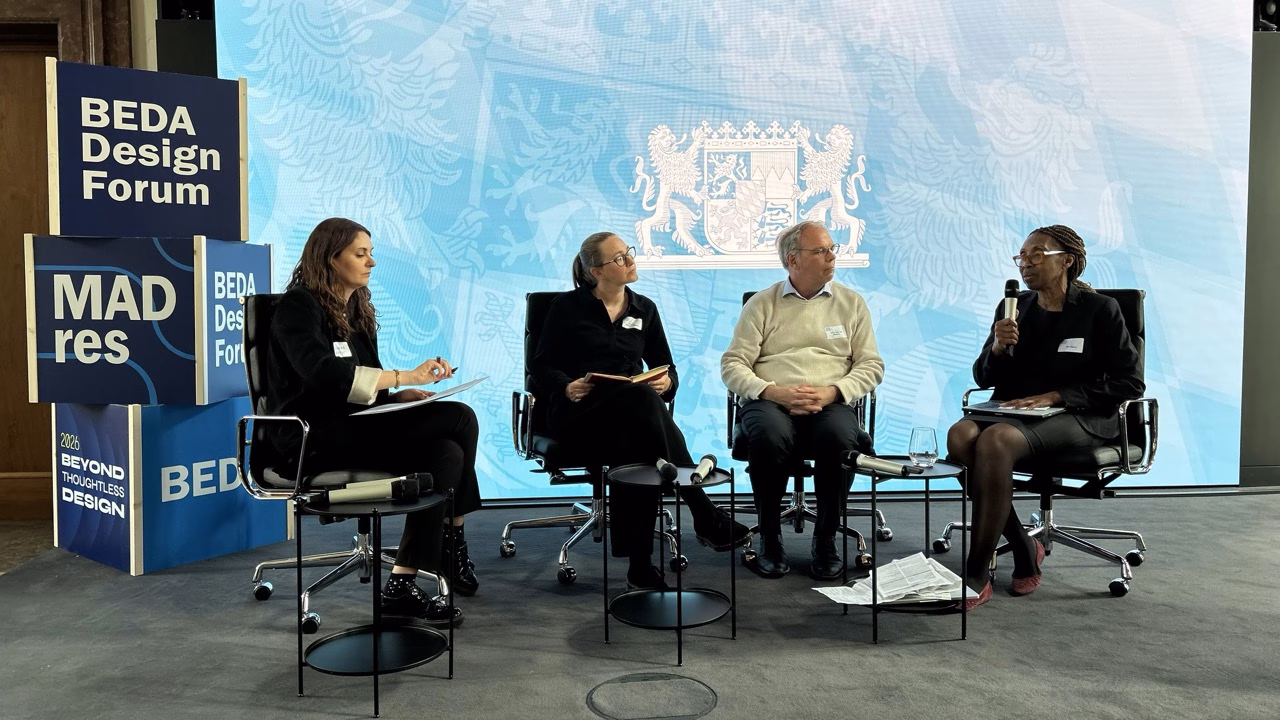 Panel discussion at the BEDA Design Forum 2026, with four speakers seated on stage in front of a large blue backdrop. One speaker is holding a microphone while the others listen and take notes. Branded displays featuring “BEDA Design Forum,” “MADres,” and the event theme “Beyond Thoughtless Design” are positioned beside the stage. Small tables with microphones and water glasses are placed in front of the panel.