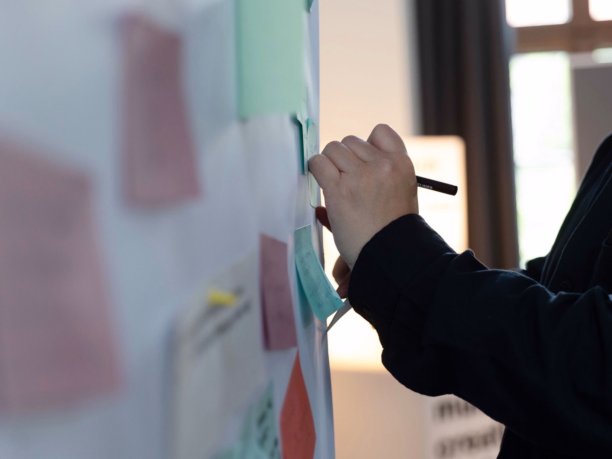 Close-up of a participant writing on a sticky note attached to a workshop board covered with colourful notes. The image focuses on a hand holding a pen, with the notes and background softly blurred, capturing a collaborative brainstorming or co-creation activity in a meeting setting.