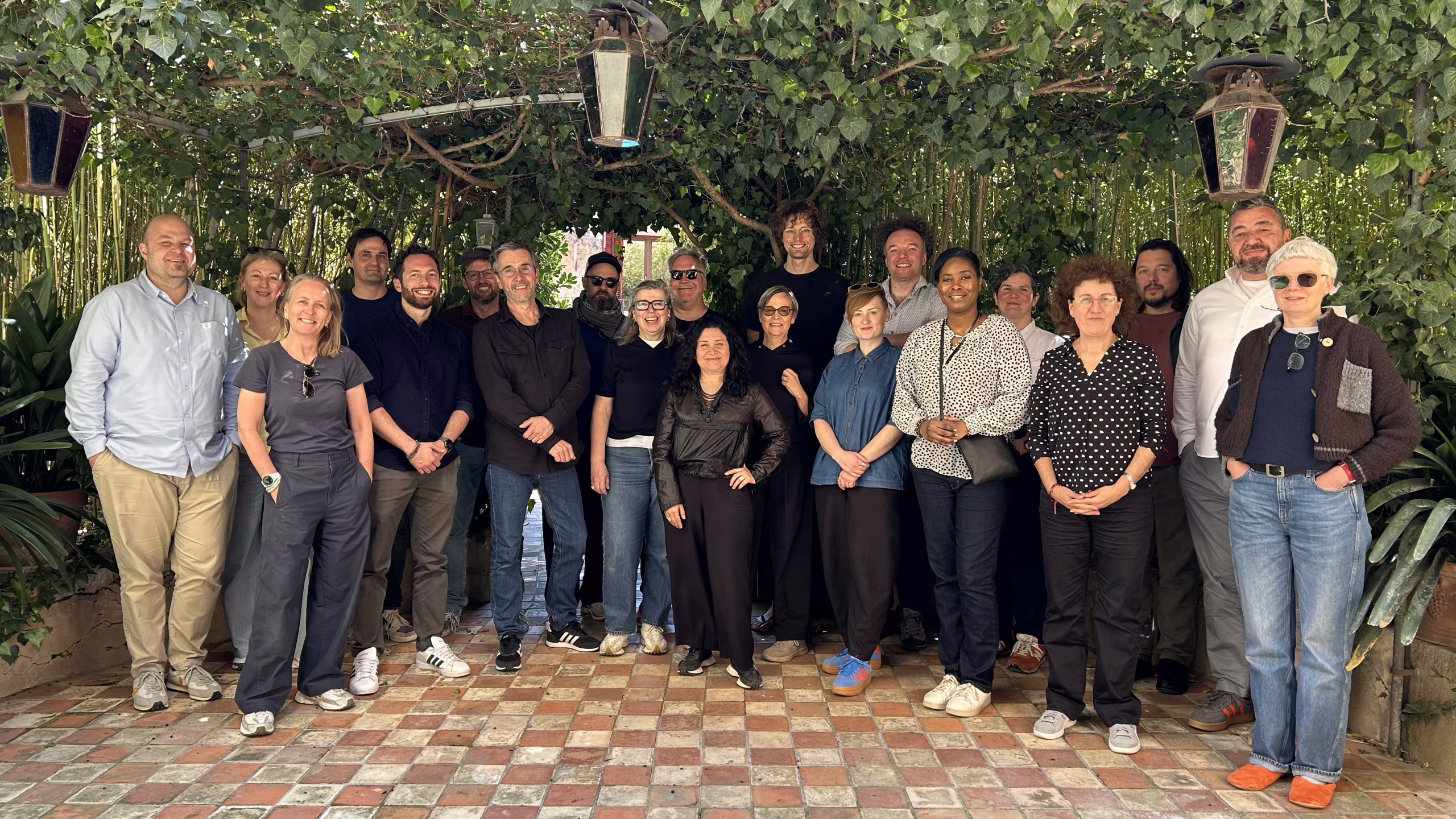 Group photo of approximately 20 people standing on a patterned tile courtyard beneath a leafy vine-covered pergola. The group is arranged in several rows under hanging lanterns, surrounded by dense greenery and bamboo. The outdoor setting is shaded, with natural light filtering through the foliage, creating an informal gathering atmosphere.